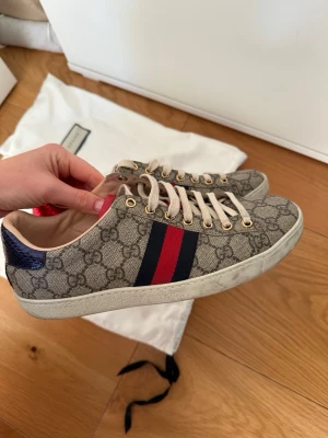 Gucci sneakers med GG-mönster, strl 39 - Säljer ett par snygga Gucci sneakers i beige canvas med klassiskt GG-mönster och röd/blå rand på sidan. Skorna har vita snören, guldfärgade öljetter och detaljer i rött och blått skinn på hälen. Insidan är ljusbeige skinn och modellen har rund tå och platt sula.