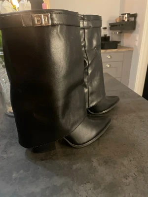 Foldover Boots, skor, stövlar - Säljer ett par svarta stövlar i slätt skinnimitation med bred, lös skaft och fyrkantigt metallspänne upptill. Stövlarna har låg klack och spetsig tå, vilket ger en cool och trendig look. Perfekta för att lyfta vilken outfit som helst.
