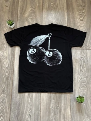Svart Stüssy t-shirt - Säljer en svart t-shirt från Stüssy i nyskick med stort vitt körsbärstryck på ryggen och klassisk Stüssy-logga på bröstet. T-shirten har rund hals och korta ärmar. Materialet är mjuk bomull som känns skön mot huden. Perfekt för dig som gillar streetwear och grafiska prints.