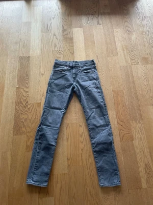 Grå Levi's slim fit jeans - Säljer ett par grå slim fit jeans från Levi's med klassisk femficksdesign och läderpatch bak i midjan. Jeansen har raka ben och är tillverkade i denim med en lätt stretch för extra komfort. Perfekta för en avslappnad och stilren look.