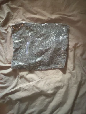 Glittrig silver tubtopp - Säljer en glittrig silver tubtopp som verkligen fångar ljuset. Perfekt för dig som vill sticka ut och glänsa. Toppen är axelbandslös och har en snygg bandeau-form. Materialet känns mjukt och följsamt mot huden.