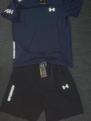 Under Armour träningsset marinblå/svart - Snyggt träningsset från Under Armour med en marinblå t-shirt och svarta shorts. Båda plaggen har logga och text i vitt. Tillverkade i ett lätt och snabbtorkande material som passar perfekt till gymmet eller löpning.