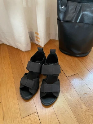 Svarta lädersandaler från Shellys London - Säljer ett par svarta sandaler från Shellys London med breda kardborreband och öppen tå. Sandalerna har chunky sula och en modern, minimalistisk look. Tillverkade i ett matt skinnmaterial som ger en stilren vibe.