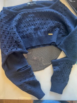 Mörkblå stickad bolero från Fratelli - Kort mörkblå stickad bolero från Fratelli med hålmönster över hela plagget. Modellen har långa ärmar och ribbstickade muddar vid ärmslut och nederkant. Perfekt att slänga över en topp för en snygg lager-på-lager-look.