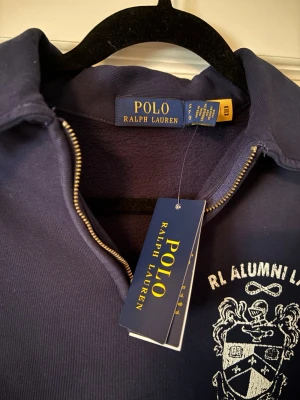 Marinblå långärmad topp Polo Ralph Lauren - Marinblå långärmad topp från Polo Ralph Lauren med vit RL Alumni LXVI-logga på bröstet. Halv dragkedja framtill och klassisk krage. Tillverkad i mjuk bomull och har en stor magficka. Perfekt för en avslappnad och stilren look.