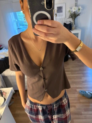 Brun v-ringad topp med knappar - Säljer en brun kortärmad topp med v-ringning och knappar framtill. Toppen har en croppad passform och är tillverkad i ett mjukt material som känns skönt mot huden. Perfekt för dig som gillar en enkel och stilren look.