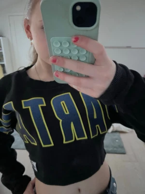 Svart croppad topp från Superdry - Cool svart croppad topp från Superdry med stora blå och gula bokstäver framtill. Ribbad rund halsringning och långa ärmar. Perfekt för dig som gillar sportig och avslappnad stil.