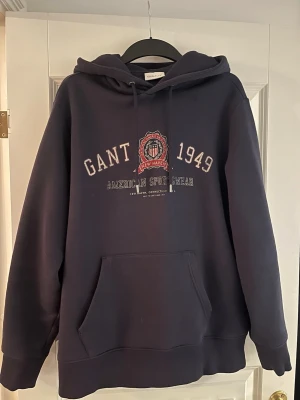 Mörkblå hoodie från GANT - Snygg mörkblå hoodie från GANT. Klassisk känguruficka och huva . Tillverkad i mjukt material. Tröjan är använd fåtal gånger och är köpt på Thernlunds för 1499kr! 
