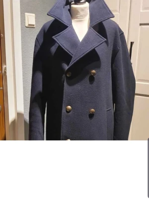 Mörkblå skepparkavaj med mässingsknappar - Stilren mörkblå skepparkavaj med bred krage och dubbla rader av mässingsknappar. Klassisk modell med två sidofickor och rak passform. Perfekt för dig som vill ha en tidlös och snygg jacka till hösten eller vintern.