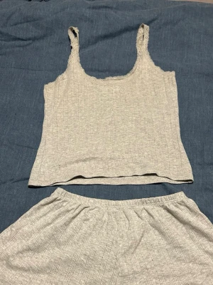 Grått ribbat set med spetsdetaljer - Mysigt tvådelat set i ljusgrått med ribbad struktur. Linnet har breda axelband med spetsdetaljer och rundad halsringning. Shortsen har resår i midjan och vågig kant nertill. Perfekt för chill dagar hemma.