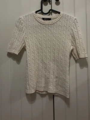 Vit kabelstickad topp från Gina Tricot - Säljer en vit kabelstickad topp från Gina Tricot med korta ärmar och rund halsringning. Tröjan har ett klassiskt kabelmönster och är gjord i mjukt material som känns skönt mot huden. 
