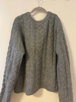 Grå kabelstickad tröja - Mysig grå kabelstickad tröja med rund hals och långa ärmar. Tröjan har ett klassiskt flätmönster och är tillverkad i ett mjukt, tjockt material. Enkel och stilren design som är lätt att matcha.