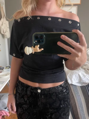 Svart offshoulder topp med nitar - Svart croppad offshoulder topp med silvriga nitar längs halsringningen och ärmslut. Toppens material känns mjukt och stretchigt, perfekt för en trendig look. Passformen är tight och toppen har korta ärmar.