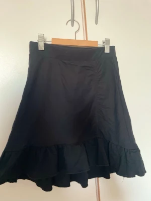 Svart volangkjol från Gina! - Säljer en svart kjol med volangkant. Kjolen har hög midja och är tillverkad i ett mjukt, lätt tyg som ger ett snyggt fall. 