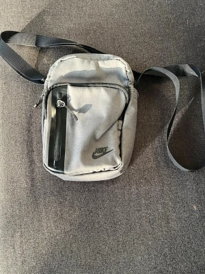 Grå axelväska från Nike - Snygg grå axelväska från Nike med svart logga på framsidan. Väskan har justerbar axelrem, ett stort huvudfack och en mindre ficka med dragkedja framtill. Tillverkad i slitstarkt tyg och perfekt för att bära det viktigaste.