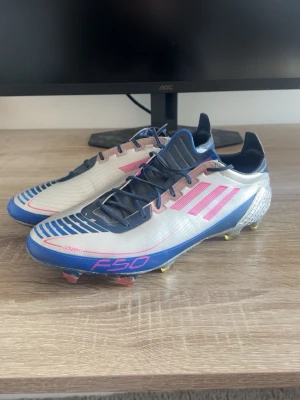 Adidas F50 Adizero Ghosted ”UCL” Limited Edition - Snygga Adidas F50 Adizero fotbollsskor i silver med rosa och blå detaljer. Skorna har klassiska tre ränder på sidan, låg profil och snörning. Yttermaterialet är syntetiskt och sulan har fasta dobbar för spel på gräs. Perfekta för dig som vill ha lätta och snabba skor på planen.