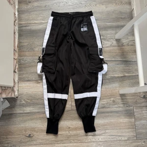 Svarta cargobyxor med vita detaljer - Säljer ett par svarta cargobyxor med breda vita ränder längs sidorna och över knäna. Byxorna har stora fickor på låren, resår i midjan med dragsko och muddar vid bensluten. Snygga detaljer med spännen och patch framtill. Perfekta för en streetwear-look.