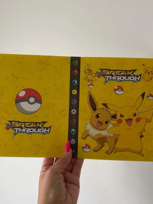 Pokémon Break Through samlarpärm - Gul samlarpärm med Pokémon Break Through-tryck, motiv av Pikachu och Eevee på framsidan samt Pokébollar och olika energisymboler. Pärmen har plastfickor inuti för att förvara och organisera samlarkort. Perfekt för Pokémonkort och andra samlarkort. Plats för 240 kort 