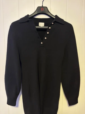 Svart stickad tröja med knappar - Klassisk svart stickad tröja med krage och fyra dekorativa knappar vid halsringningen. Tröjan har långa ärmar och ribbade muddar, perfekt för en stilren och enkel look. Passar till det mesta och är superenkel att matcha.