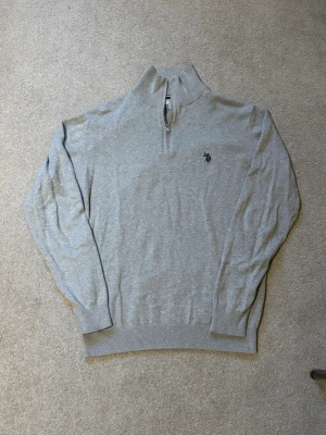 Quarterzip U.S. Polo Assn. - Snygg ljusgrå tröja med halv dragkedja från U.S. Polo Assn. Broderad logga på bröstet och ribbade muddar vid ärmslut och nederkant. Perfekt för lager på lager och enkel att matcha med jeans eller chinos.