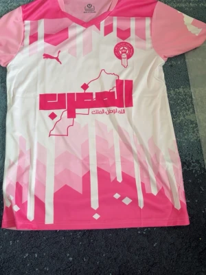 Marocko matchtröja rosa/vit Puma M - Snygg Marocko matchtröja från Puma i rosa och vitt med grafiskt mönster och tryck på bröstet. Tröjan har korta ärmar, V-ringning och officiellt förbundsmärke. Tillverkad i lätt och ventilerande material, perfekt för fotboll eller träning.