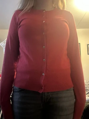 Röd kofta med guldknappar - Snygg röd kofta med rund halsringning och långa ärmar. Koftan har ribbade muddar och knäpps med guldfärgade knappar framtill. Materialet är mjukt och stickat, perfekt för lager på lager.