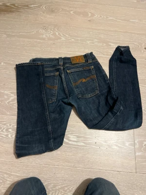 Mörkblå jeans från Nudie Jeans - Säljer ett par mörkblå jeans från Nudie Jeans med klassisk femficksmodell och kontrastsömmar i orange. Jeansen har en snygg broderad detalj på bakfickorna och patch med logga i läder baktill. Perfekta för en avslappnad och stilren look.