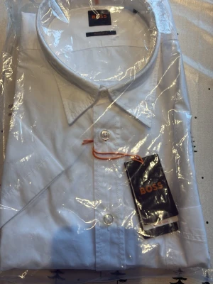 Vit skjorta från BOSS med klassisk krage - Snygg vit skjorta från BOSS med klassisk krage och knappar framtill. Tillverkad i bomull för en skön och stilren känsla. Perfekt för dig som vill ha en clean och tidlös look. Diskret BOSS-logga på insidan av kragen och etikett kvar. Skriv för minsta lilla fråga.