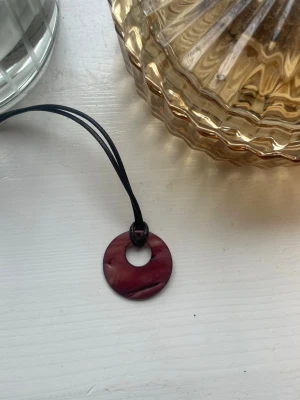 Runt halsband med lila pärlemorhänge - Unikt halsband med ett runt, lila pärlemorhänge i donut-form. Hänget har en skimrande yta med naturliga färgskiftningar och sitter på ett svart snöre. Perfekt för dig som gillar boho eller retrostil.