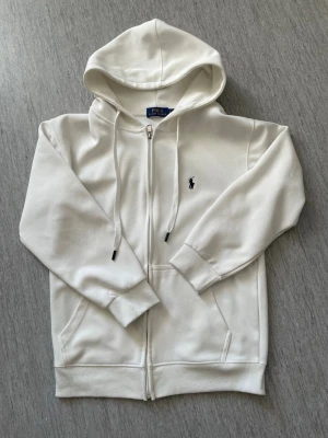 Vit hoodie från Polo Ralph Lauren - Snygg vit hoodie med dragkedja från Polo Ralph Lauren. Klassisk design med huva och dragsnören, två fickor framtill och den ikoniska svarta loggan broderad på bröstet. 