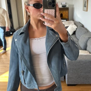 Blå mocka skinnjacka med dragkedja - Snygg blå skinnjacka i mocka med silverfärgade dragkedjor och knappar. Jackan har klassisk bikerstil med bred krage och asymmetrisk dragkedja framtill. Perfekt för dig som vill ha en cool och trendig look.