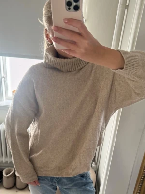 Beige polotröja i stickat material - En stilren beige stickad polotröja med bred krage och långa ärmar. 