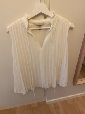 Vit ärmlös blus - Snygg vit blus med knytning  Blusen är ärmlös och har en liten ståkrage med knytband. Perfekt för en stilren och luftig look. Passar till både jeans och kjol.
