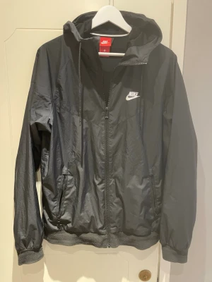 Svart vindjacka från Nike med huva - Snygg svart vindjacka från Nike med vit logga på bröstet. Jackan har dragkedja framtill, två fickor med dragkedja och en praktisk huva. Tillverkad i lätt och tåligt material, perfekt för blåsiga dagar. Resår vid ärmslut och nederkant för skön passform.