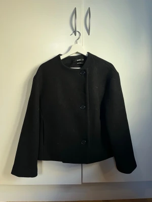 Svart kappa - Minimalistisk svart kappa från bikbok. Rak, boxig passform med rund halsringning och tre stora knappar framtill. Perfekt för dig som gillar stilren design och klassiska plagg.