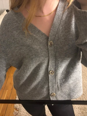 Grå kofta  - Mysig grå kofta med stora pärlemorknappar och v-ringning. Koftan har långa ärmar och är tillverkad i ett mjukt stickat material. Jätteskön och i väldigt bra skick.