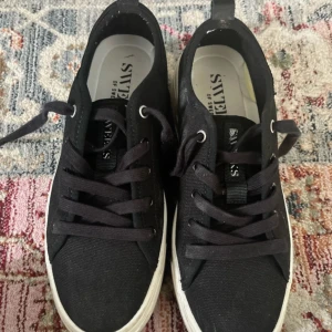 Svarta sneakers från Sweeks of Sweden storlek 37  - Storlek 37. Svarta sneakers från Sweeks of Sweden med klassisk design. Skorna har svart textilöverdel, vita gummisulor och svarta snören. Rund tå och platt sula ger en clean och stilren look som passar till det mesta. Perfekta för dig som gillar enkel och tidlös stil. De har en liten fläck på ena skon (se bild 4) men inget som stör! Använd 4-5 gånger. 