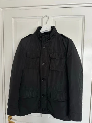 Field dunjacka från MooRER - Stilren svart dunjacka från MooRER med hög krage, fyra fickor framtill och quiltat foder i brunt. Jackan har knäppning med både dragkedja och knappar samt detaljer som axelklaffar och innerfickor. Perfekt för vinterdagar och vår. Nypris 17000