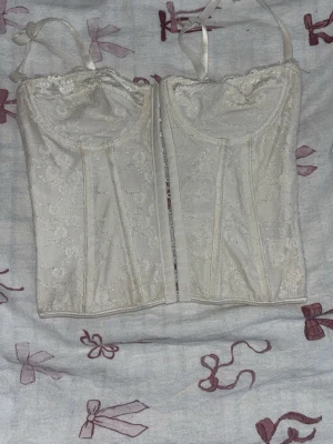 Vit spetskorsett med snörning - Snygg vit korsett med spetsdetaljer och justerbara axelband. Framsidan har hyskknäppning och baksidan har dekorativ snörning. Materialet är spets och satinliknande tyg som ger en lyxig känsla. Perfekt för att skapa en feminin siluett. Använt några enstaka gånger men inte mer än så.