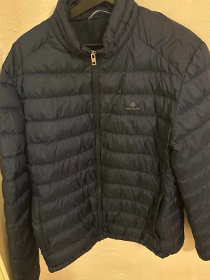 Mörkblå pufferjacka från Gant - Snygg mörkblå pufferjacka från Gant med diskret logga på bröstet. Jackan har dragkedja framtill, ståkrage och quiltad design med horisontella sömmar. Perfekt för kalla dagar och tillverkad i lätt, vadderat material.