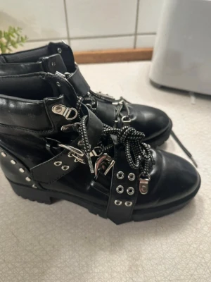Svarta boots med nitar och spännen - Säljer ett par svarta kängor i skinnimitation med grov sula, snörning och coola detaljer som nitar, spännen och metallhakar. Skosnörena är svarta med vita detaljer och kängorna har en edgy look som passar till streetstyle.