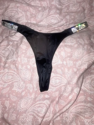 Svart stringtrosa med strassband - Snygg svart stringtrosa med glittrande strassdetaljer på sidorna. Trosan har en smal passform och är tillverkad i ett glansigt, stretchigt material som känns mjukt mot huden. Perfekt för dig som gillar att sticka ut med lite extra bling. Strl M men passar som S, använt en gång bara.