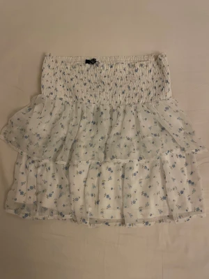 Vit blommig volangkjol från Vero Moda - Supersöt kort kjol från Vero Moda i vitt med små blå blommor. Kjolen har smockad midja och dubbla volanger som ger en luftig och romantisk känsla. Perfekt för varma dagar och enkel att matcha med en enkel topp.