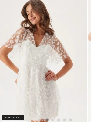 Vit kortklänning med blommig spets - Superfin vit kortklänning med blommig spets och v-ringad hals. Klänningen har korta ärmar i transparent spets och ett sött blommigt mönster över hela. Perfekt för studenten. Aldrig använd endast testad.