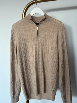 Beige kabelstickad tröja Sand - Kabelstickad beige tröja från Sand Copenhagen med halv dragkedja vid halsen. Tröjan har ribbade muddar och hög krage, perfekt för lager på lager. Materialet känns mjukt och exklusivt, och färgen är lätt att matcha till olika outfits. Står S men skulle säga att den är mer av en M. Helt ny då den inte har används sen något sen den köptes.