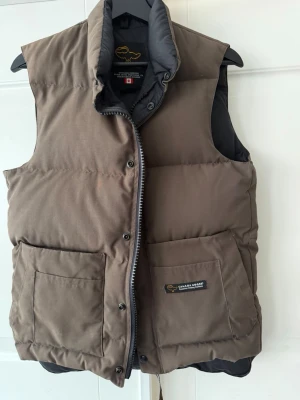 Brun dunväst  - Säljer en brun dunväst med hög krage och två stora framfickor. Västen har dragkedja och tryckknappar framtill samt diskret logga på ena fickan. 