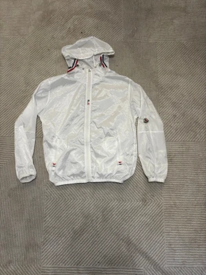 Vit  vindjacka - Snygg vit vindjacka från  med huva och dragkedja framtill. Jackan har elastiska muddar, två fickor med dragkedja och M logga på ärmen. Detaljer i rött, vitt och blått vid fickor och huva. Ni vet varför priset är som det är. Skicka gärna prisförslag eller frågor.😊