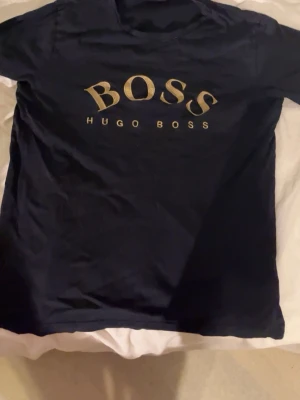 Svart t-shirt från Hugo Boss - Snygg svart t-shirt från Hugo Boss med guldfärgad logga tryckt på bröstet. Klassisk rund halsringning och korta ärmar. Perfekt för dig som vill ha en stilren och trendig look med exklusiv känsla.