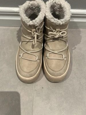 Beige fejk Inukiis med fuskpäls - Säljer ett par beige fejk Inukiis med mysigt fuskpälsfoder och kraftig sula. Skorna har snörning framtill och är perfekta för kalla dagar. De har en klassisk design och är riktigt sköna att ha på sig.