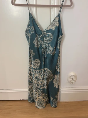 Blå mönstrad klänning med snörning - Populär,  snygg blå klänning med vitt blommigt mönster och smala axelband. Klänningen har v-ringning och snörningsdetalj framtill. Lätt och luftig modell som passar perfekt till varma dagar.